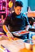 Confident focused male cook in black uniform and gloves placing freshly cut salmon meat in tray for preparing meal at illuminated sushi bar