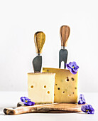 Cheese with cheese knifes on wooden cutting board with blue flowers at white background. Delicious aromatic mature hard cheese. Front view.