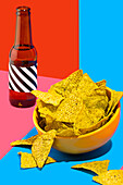 Ein lebhaftes Stillleben mit einer Schüssel knuspriger Nacho-Chips neben einer dunklen Flasche vor einem geteilten Hintergrund aus Rot und Blau