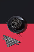 Top view of black spaghetti in a bowl with chopsticks placed on black and red background near napkin and fork