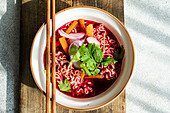 Top view of bowl with beetroot soup with onion, coriander and noodles in Asian style served on bowl and chopsticks on wooden cutting board against gray surface in daylight