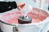 Nahaufnahme einer abgeschnittenen, nicht erkennbaren Hand, die Zucker in eine Zuckerwattemaschine gießt, in der sich rosa Zuckerwatte bildet
