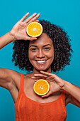 Optimistic young Hispanic female with curly black hair smiling and looking at camera while holding orange slices against blue background