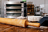 Closeup of wooden rolling pin with flour in bowl placed on table with large oven and kitchen utensils in background at bakehouse