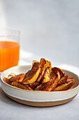 Fried potato with paprika spice served on ceramic plate near orange juice against gray background
