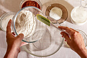 Top view of hands of an anonymous crop person sifting flour into a glass bowl with baking ingredients around on a table