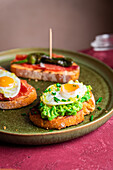 Delicious homemade Spanish tapas toasts with fried eggs, avocado and ham served on round plate near crispy bread with bacon on table