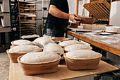 Seitenansicht eines fokussierten anonymen männlichen Bäckers, der ungekochtes Sauerteigbrot in Formen mit Plastik auf einem Holztisch in der Küche einer Bäckerei abdeckt