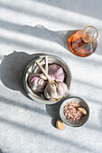 Draufsicht auf frische rohe Knoblauchzwiebeln in einer Keramikschüssel, begleitet von grobem Salz und einer Braunglasflasche, alles vor einem strukturierten hellgrauen Hintergrund