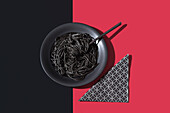 Top view of black spaghetti in a bowl with fork placed on black and red background near napkin