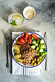 Top view of healthy lunch bowl with boiled organic buckwheat, fresh cucumber and tomato, and fermented tomato and olives served with glass of pure water with lemon, ice and mint against gray surface