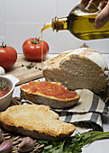 Crop hand of anonymous person pouring olive oil from bottle on wholegrain bread with tomato spread between unpeeled garlic and chopping board on table