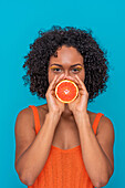 Happy young Hispanic female with curly hair covering mouth with slice of orange and looking at camera against blue background