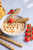 Hochformatiger gesunder Pflanzenteller mit Hummus und Tomaten, serviert mit Brotstäbchen in einer Schale neben einer Serviette vor unscharfem Hintergrund