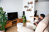 Blick von oben auf einen zufriedenen jungen Afroamerikaner in Freizeitkleidung, der mit einer Schüssel Popcorn sitzt und einen Fernseher mit schwarzem Bildschirm einschaltet, während er sich zu Hause auf einem bequemen Sofa ausruht