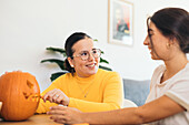 Happy women sitting at table carving with tool scary teeth on orange pumpkin and preparing for Halloween decoration in day