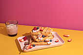 From above tasty sweet waffles topped with berries fruits sauce and cream served on pink tray on colorful table background near glass with hot milk beverage in light kitchen