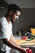 Side view of young ethnic African American male in casual t shirt washing fresh bell pepper in sink with water stream while cooking healthy salad in kitchen