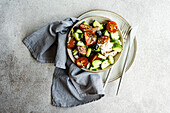 Draufsicht auf Keramikschüssel mit leckerem gesundem Salat und Gemüse mit Gurken- und Tomatenscheiben, Besteck und Stoff auf grauem Tisch