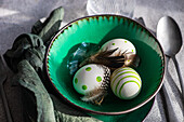 High angle closeup of Easter table setting, showcasing a vibrant green ceramic plate with two decorative Easter eggs adorned with white and green patterns and delicate feathers, placed on gray surface between napkin and cutlery and glass of water