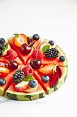 High angle of ingredients consisting of strawberry apple raspberry grapes blackberry plum blueberry peach placed on round cut slices on white surface while preparing watermelon pizza