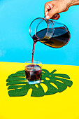 Cropped hand of anonymous person pouring juice from jug in glass on leaf decor on yellow backdrop against blue background