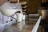 Anonymous person pouring fresh milk from a jug into a glass on a counter with a bakery background
