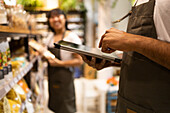 Cropped unrecognizable African American male seller in apron standing near shelves with groceries and looking at screen of tablet while checking information