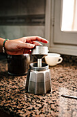 Nahaufnahme einer anonymen Hand, die eine italienische Mokkakanne auf einer Küchentheke zusammensetzt, mit unscharfem Küchengeschirr im Hintergrund, das auf die Zubereitung von Kaffee zu Hause hinweist