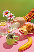Unrecognizable person pouring fresh milk into cup of hot aromatic coffee placed on table with delicious croissants and banana decorated by flowers in vase