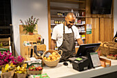 Happy young African American male seller in apron smiling and entering information in computer while standing at counter during work