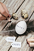 Eine Person stempelt das Wort "Ostern" auf ein weißes Ei, das mit Wachteleiern, Federn und Anhängern, auf denen "Happy" und "Eier" steht, auf rustikalem Holz liegt