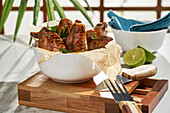 Tasty grilled marinated chicken wings placed in bowl and served with sliced lime on wooden cutting board with knife on table against blurred background in daylight