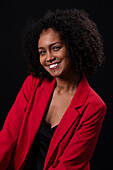 Portrait of smiling young Hispanic female model with Afro hairstyle in red coat looking at camera while standing against black background