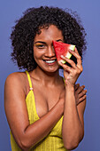Friendly charismatic African American female model smiling and looking at camera while hiding eye behind slice of sweet watermelon