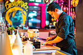 Side view of confident male cook with concentration holding knife and cutting nori seaweed on chopping board at illuminated counter in sushi bar