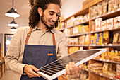 Positive seller male in casual clothes and apron smiling while standing near shelves holding and looking at portable solar panel in modern supermarket