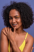 Positive Hispanic young female with curly black hair smiling and touching chin while looking at camera on blue background