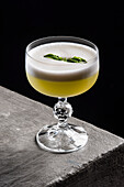 High angle of refreshing alcoholic pisco sour cocktail with froth and mint served in glass placed on gray stone table