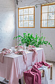 Wooden table with pink tablecloth and dishware placed near bench covered with cloth against potted plant and white brick wall