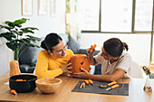 Focused young Hispanic female friends looking down while sitting at table with Halloween kit bowls and carving eye with sharp tool on orange pumpkin