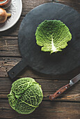 Whole raw savoy cabbage and cabbage leaf on black cutting board on rustic wooden kitchen table with knife. Preparing food with seasonal autumn vegetable. Top view