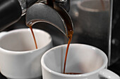 Professional coffee maker pouring hot drink into in two white cups on blurred background