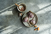 From above of raw garlic bulbs with a bowl of coarse salt on a textured grey surface illuminated by soft sunlight casting shadows