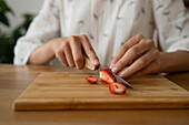 Crop anonymous female in white cloth sitting and cutting red strawberry on board over wooden table while making strawberry jam indoors