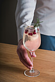 A hand presenting a rosemary winter cocktail in a stemmed glass, garnished with cranberries and a sprig of rosemary, against a neutral background.
