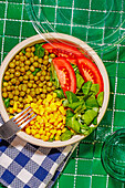 Top view of salad bowl with slices of tomato, spinach leaves, corn kernels and peas placed on napkin on green surface with fork near glass of water