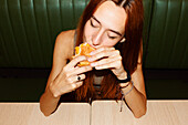 High angle of young woman with red hair and piercing with closed eyes sitting at table and enjoy eating hamburger in restaurant