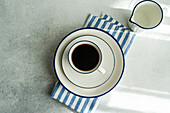 Draufsicht auf einen frisch gebrühten Tropfkaffee in einer weißen Tasse auf einer Untertasse mit blauen Streifen, daneben ein Milchkännchen und eine gestreifte Serviette