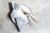 Top view of minimalistic rustic table setting with white plate cutlery and striped napkin on gray surface in daylight